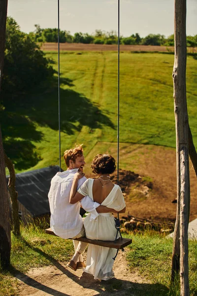 Marié rousse excité embrassant mariée en robe blanche tout en s'amusant sur swing dans un cadre rural — Photo de stock