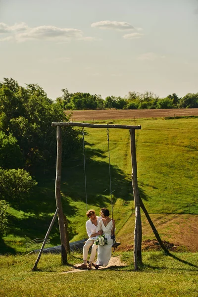 Happy and stylish interracial newlyweds on swing in countryside with scenic landscape, tranquility — Stock Photo