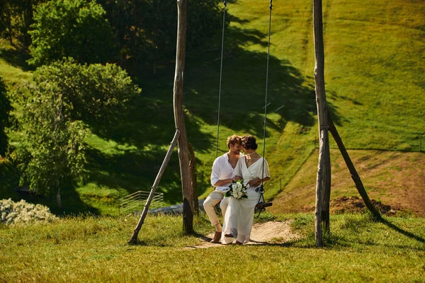Feliz momento de pareja multiétnica recién casada en traje de estilo boho en swing en el paisaje - foto de stock