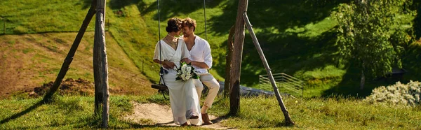 Joyeux interracial jeunes mariés en tenue de style boho sur swing dans un cadre rural pittoresque, bannière — Photo de stock