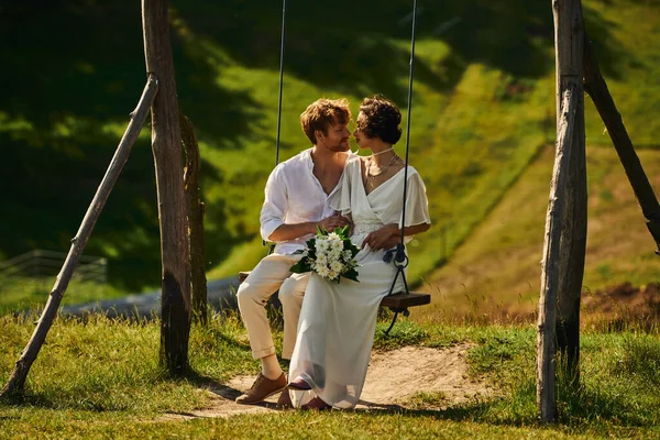Matrimonio rustico, sposo rossa e sposa asiatica con bouquet guardando l'un l'altro su swing — Foto stock