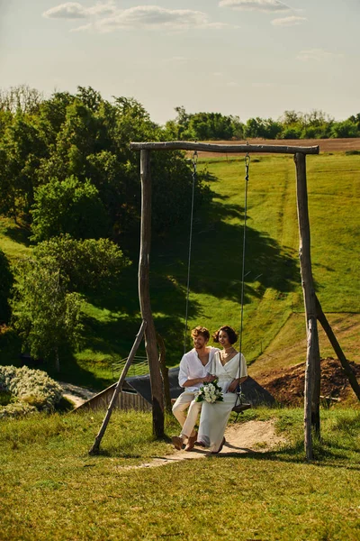 Jeunes mariés multiethniques insouciants et élégants sur le swing rustique dans la campagne avec un paysage pittoresque — Photo de stock