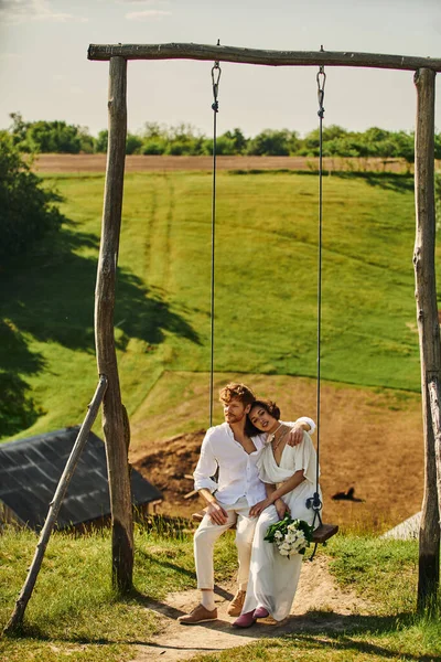 Redhead man embracing elegant asian bride with flowers on swing in rural setting, rustic wedding — Stock Photo