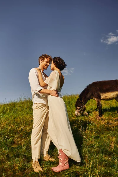 Feliz ruiva homem abraçando jovem asiático noiva perto burro pastando no verde prado, rústico casamento — Fotografia de Stock