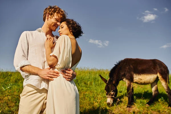 Jovem asiático homem abraçando encantador asiático noiva no branco vestido perto burro pastando no prado — Fotografia de Stock