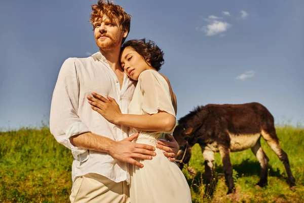 Just married interracial couple in boho style attire embracing near donkey grazing on background — Stock Photo