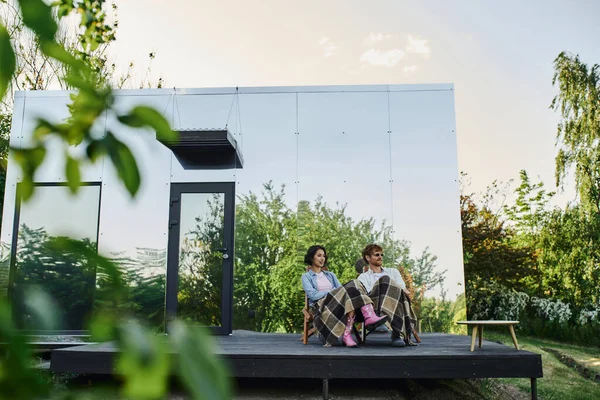Молода багатоетнічна пара насолоджується свіжим повітрям, сидячи під ковдрами на ганку скляного будинку — стокове фото
