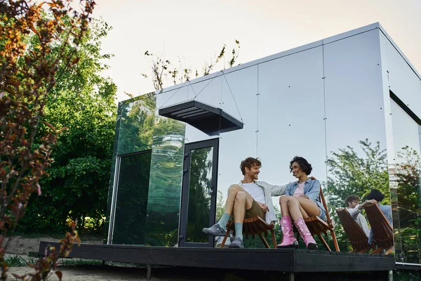 Счастливая многонациональная пара, сидящая на стульях и глядя друг на друга возле современного стеклянного дома — стоковое фото