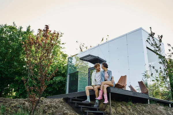Беззаботная межрасовая пара сидит на крыльце современного травяного домика и улыбается друг другу — стоковое фото