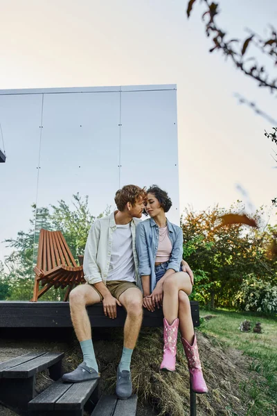 Multiethnisches Paar in stilvoller Freizeitkleidung küsst sich auf der Veranda eines Glashauses auf dem Land — Stockfoto