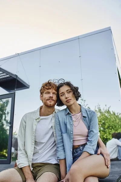 Verträumtes multiethnisches Paar lächelnd und wegschauend in der Nähe von Glashaus auf dem Land, Ruhe — Stockfoto