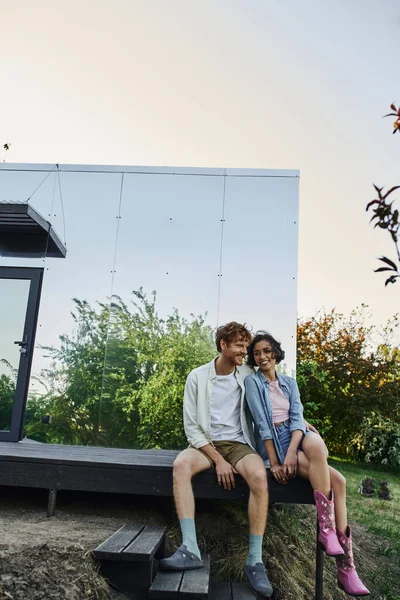 Радісна багатоетнічна пара сидить на ганку сучасного скляного будинку у вихідні в сільській місцевості — стокове фото