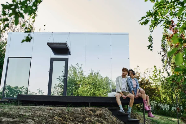 Cheerful interracial couple sitting on porch of modern glass house in countryside setting — Stock Photo