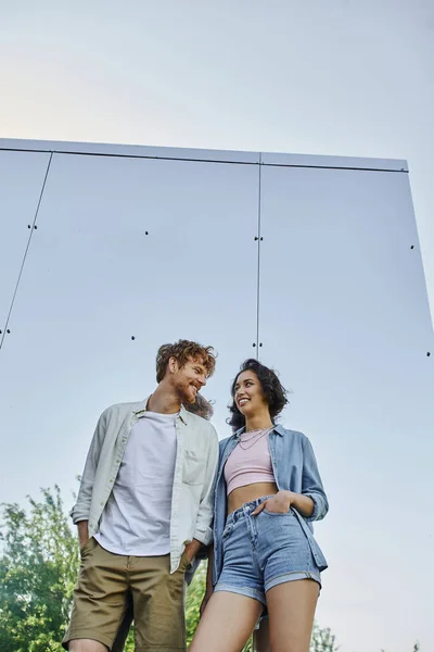 Interracial couple with hands in pockets smiling at each other near mirror wall of glass house — Stock Photo