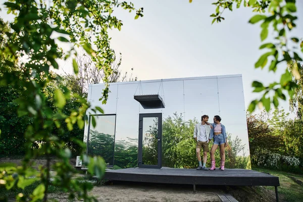 Полная длина счастливой и стильной межрасовой пары, стоящей на крыльце стеклянного дома в сельской местности — стоковое фото