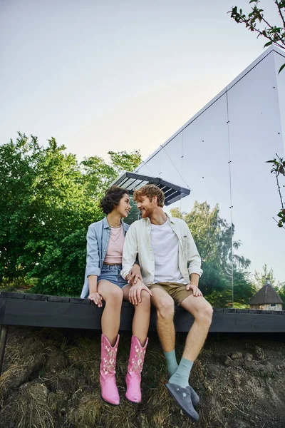 Fröhliche multiethnische Paar sitzt auf Veranda in der Nähe von Glashaus und schauen einander an, Glück — Stockfoto