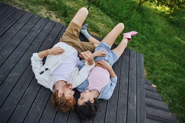 Carefree multiethnic couple in stylish casual attire lying down on wooden porch outdoors, top view — Stock Photo