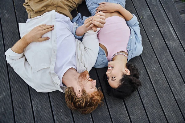 Joyful interracial couple lying down on wooden porch and looking at each other outdoors, top view — Stock Photo
