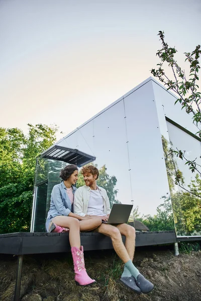 Feliz pareja interracial con portátil sentado en el porche cerca de la moderna casa de cristal en el campo - foto de stock