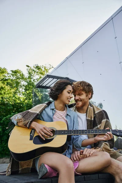 Feliz mulher asiática tocando guitarra acústica para namorado na varanda perto de casa de vidro no campo — Fotografia de Stock