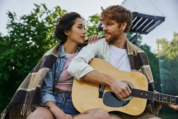 Young redhead man playing acoustic guitar to asian girlfriend near glass house, countryside date — Stock Photo