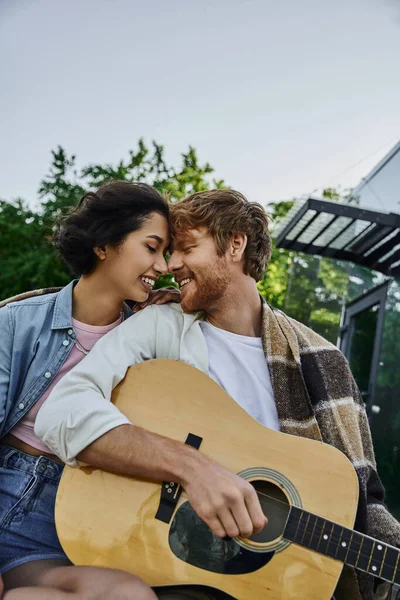 Alegre pelirroja hombre jugando acústica guitarra a joven asiática novia cerca de vidrio casa en campo - foto de stock