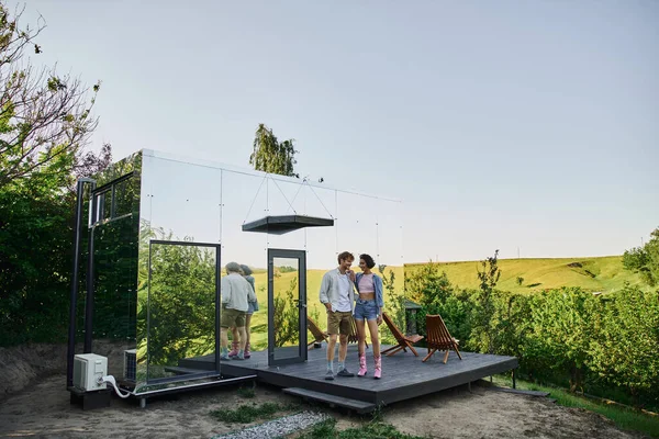 Toute la longueur de couple interracial élégant debout sur le porche de la maison en verre moderne dans la campagne — Photo de stock