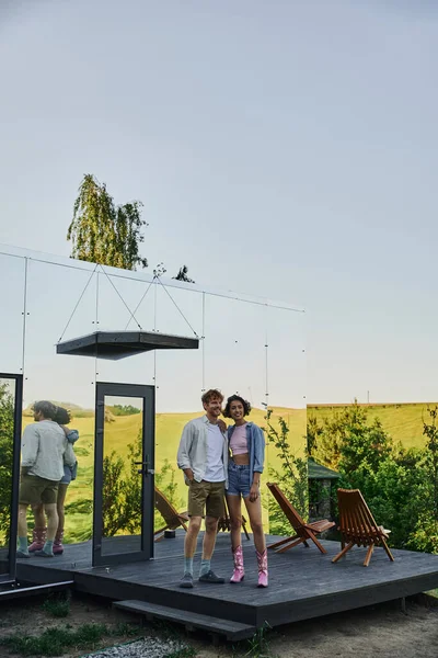 Comprimento total do feliz casal interracial olhando para longe no alpendre da moderna casa de vidro no campo — Fotografia de Stock
