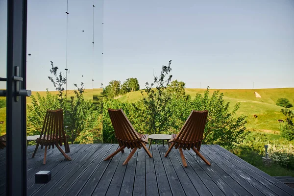 Holzstühle und Couchtisch auf Veranda in der Nähe modernes Glashaus mit Blick auf malerische Landschaft — Stockfoto