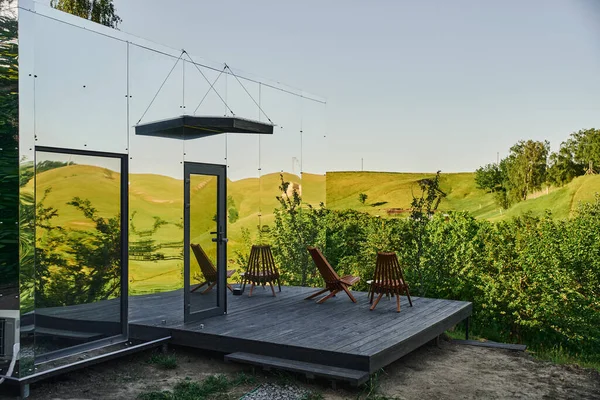 Екологічно чистий скляний будинок зі стільцями на дерев'яному ганку і видом на мальовничий сільський пейзаж — стокове фото