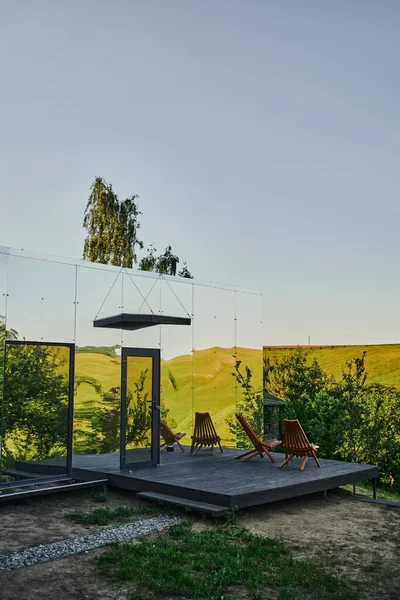 Современный стеклянный дом с деревянным крыльцом возле зеленых холмов под голубым небом в живописной сельской местности — стоковое фото