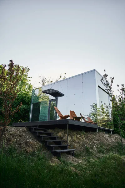 Modernes umweltfreundliches Glashaus mit Holztreppe und Stuhl auf Veranda, ruhige Landschaft — Stockfoto