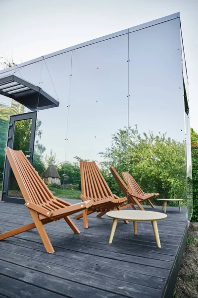 Silla de madera y mesa de centro cerca de la moderna casa de vidrio ecológico en un entorno rural tranquilo - foto de stock