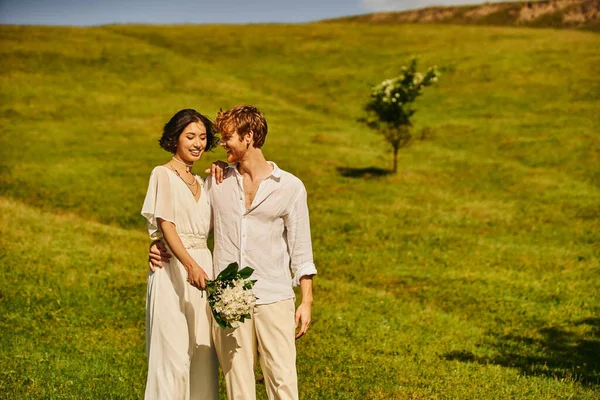 Cheerful redhead man embracing asian woman with wedding bouquet on green meadow in countryside — Stock Photo