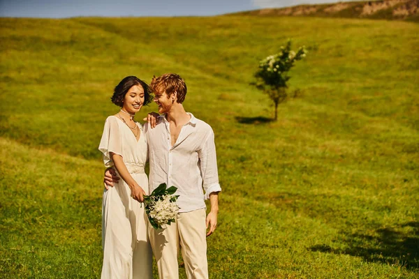 Alegre pelirroja hombre abrazando asiático mujer con boda bouquet en verde prado en campo - foto de stock