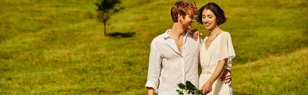 Alegre pelirroja novio abrazando asiático novia con boda ramo en paisaje campo, bandera - foto de stock