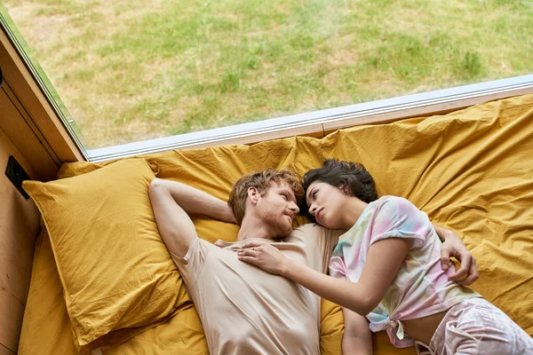 Vista dall'alto di rossa uomo abbracciare ragazza asiatica mentre sdraiati insieme su biancheria da letto gialla a casa — Foto stock