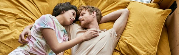 Vista superior do casal interracial deitado na cama amarela e sorrindo um para o outro em casa, banner — Fotografia de Stock