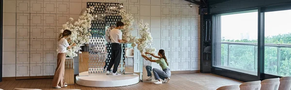 Творческий коллектив, организующий праздничный декор с белыми цветущими цветами в современном зале мероприятий, баннер — стоковое фото