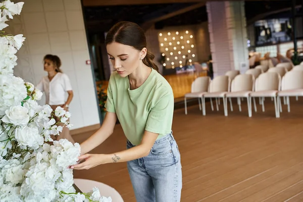 Young tattooed woman working with blooming floral composition in event hall near blurred colleague — Stock Photo