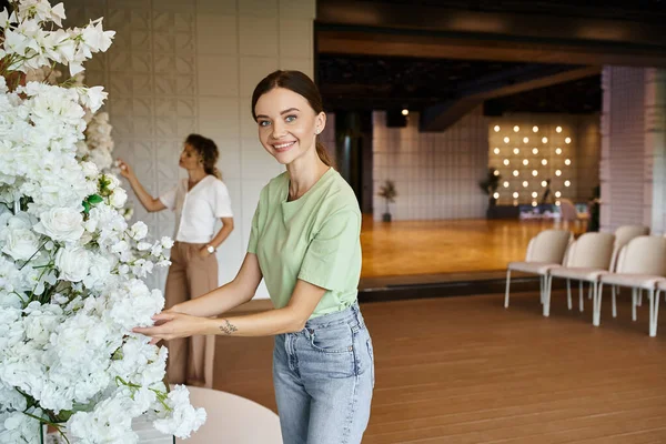 Glücklicher Dekorateur blickt in die Kamera, während er in der Veranstaltungshalle Blumenmuster in der Nähe verschwommener Kollegen arrangiert — Stockfoto