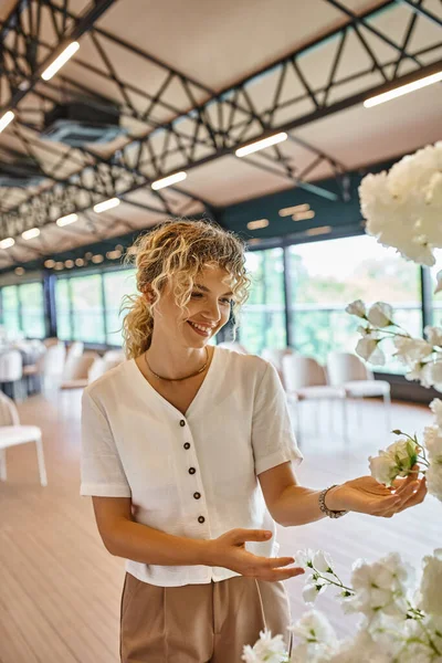 Mujer rubia alegre con el pelo ondulado sonriendo cerca de flores blancas en la sala de eventos, florista creativa - foto de stock