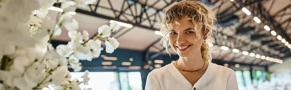 Felice donna bionda sorridente vicino composizione floreale bianca in sala eventi, fiorista al lavoro, banner — Foto stock