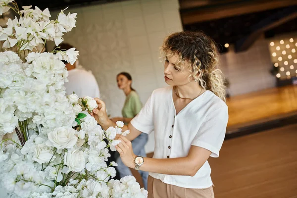 Mujer alegre mirando la cámara cerca de la decoración floral y colega en el fondo borroso en la sala de eventos - foto de stock