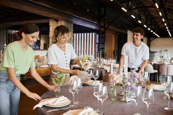 Радостная команда, организующая праздничную сервировку стола и возлагающая цветы возле посуды в зале мероприятий — стоковое фото