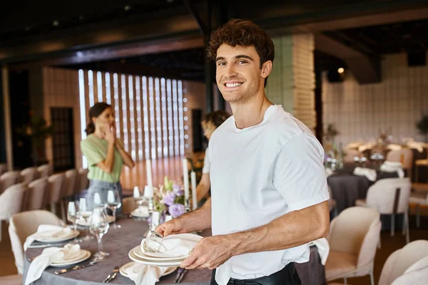Smiling event manager with plates and napkins looking at camera near colleagues and festive table — Stock Photo