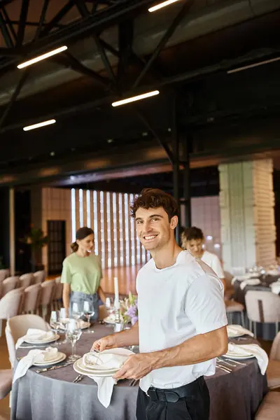 Fröhlicher Eventdekorateur mit Tellern und Servietten, der in die Kamera neben festlichem Tisch und Kollegen blickt — Stockfoto