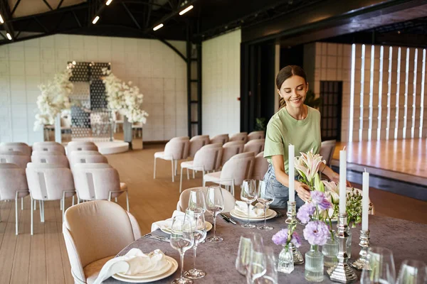 Felice event manager sorridente vicino al tavolo con cornice festosa, fiori e candele nella sala eventi — Foto stock