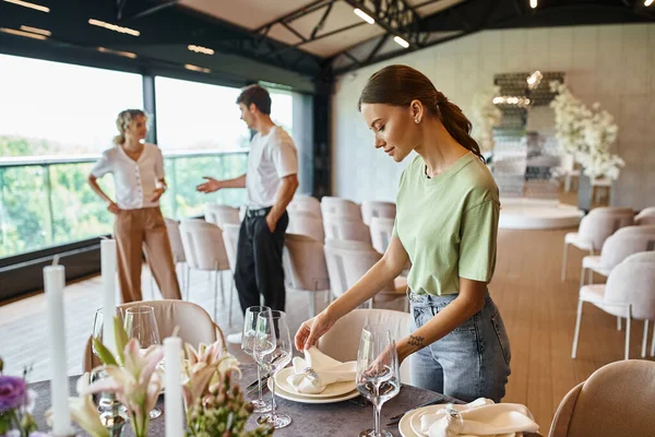 Young decorator arranging banquet table setting near colleagues talking in celebration hall — Stock Photo