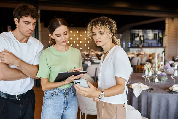 Young woman writing notes on clipboard near colleague and team lead in event hall, banquet setup — Stock Photo